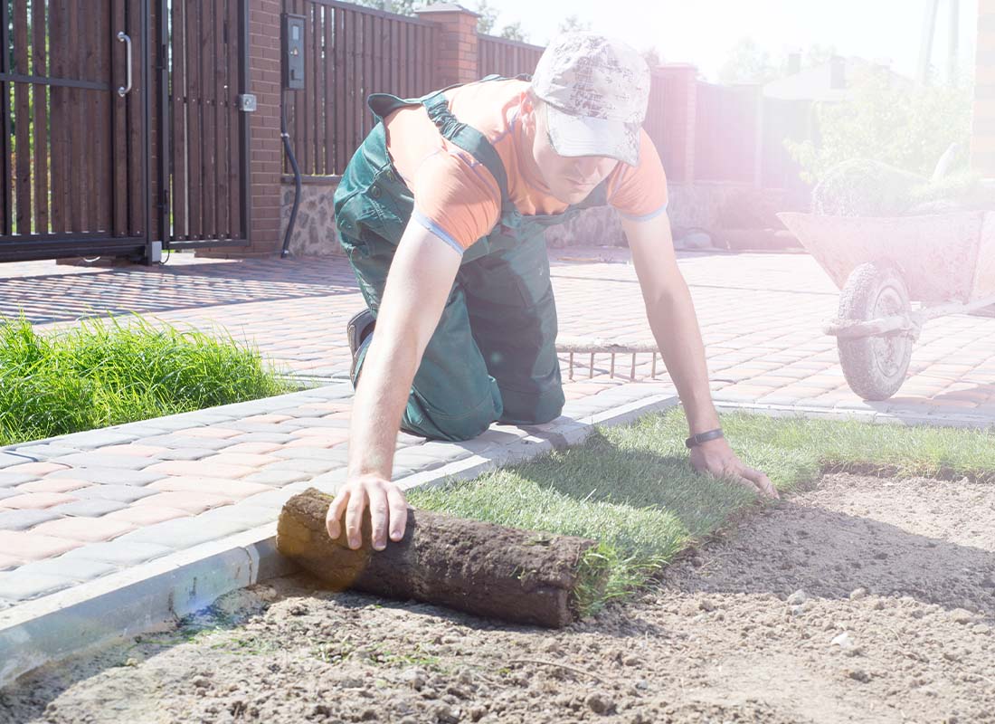 Landscaping Services Insurance - Landscape Gardener Laying Turf for New Lawn with Wheelbarrow of a Backyard Patio in a Fenced in Yard on a Bright Sunny Day